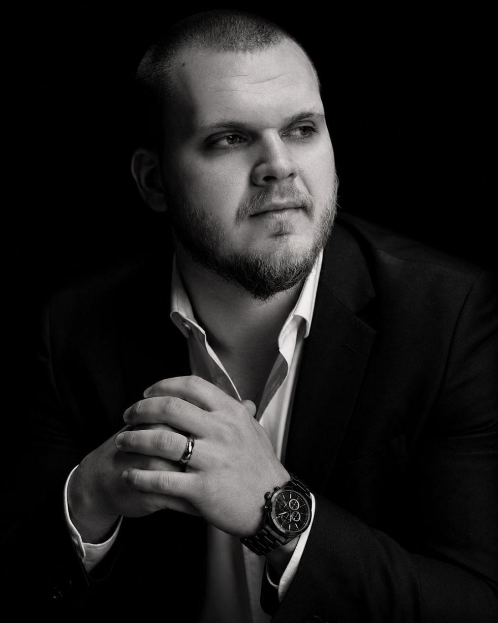 Black and white portrait of Isaac Mashman, seated, hands clasped, wearing a suit with an open collar.